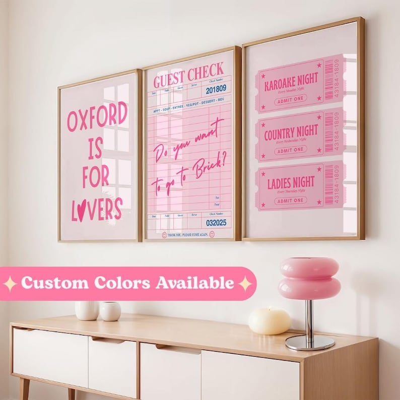 May include: Three framed pink art prints with gold frames. The prints feature text: "OXFORD IS FOR LOVERS", a "GUEST CHECK" design, and three "NIGHT" tickets. A pink lamp and decor sit on a wooden cabinet.