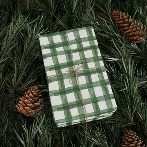 May include: A rectangular gift box wrapped in white paper with a green watercolor plaid pattern, tied with thin brown twine in a simple bow. The box is positioned among green pine needles and several brown pine cones.