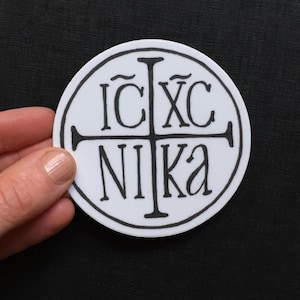 May include: A white round sticker with a black cross and the text "IC XC NIKA" in a stylized font.
