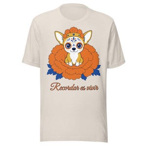 Peut inclure: Un t-shirt beige à manches courtes orné d'un graphique représentant un petit chien au maquillage de tête de mort, portant une couronne de fleurs orange, assis dans une grande fleur orange avec des feuilles bleues. Le texte Recordar es vivir est imprimé en dessous.