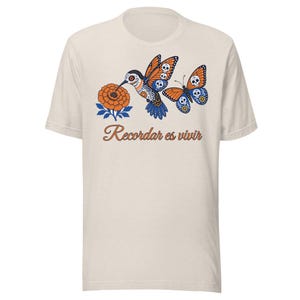 Peut inclure: Un t-shirt couleur crème arborant un motif composé d'une fleur de souci orange, d'un colibri et d'un papillon, tous ornés de motifs de crânes. Sous le dessin, le texte "Recordar es vivir" est imprimé en police cursive orange.