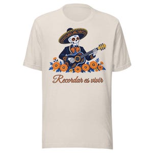 Peut inclure: Un t-shirt couleur crème avec un motif représentant un squelette portant un chapeau et un costume de mariachi tout en jouant de la guitare. Sous le squelette se trouvent des œillets d'Inde orange et le texte Recordar es vivir.