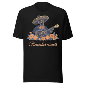 Peut inclure: Un t-shirt noir avec un motif représentant un squelette en tenue de mariachi et sombrero, jouant de la guitare. Le squelette est entouré de fleurs orange. Sous le motif, le texte indique, Recordar es vivir.
