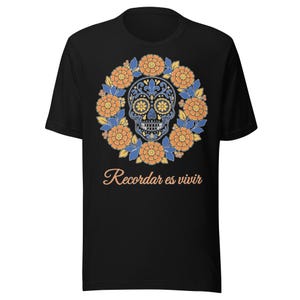 Peut inclure: Un t-shirt noir à manches courtes arborant un motif central représentant un crâne bleu et blanc entouré d'une couronne circulaire de fleurs de souci orange et de feuilles bleues, avec le texte Recordar es vivir imprimé en orange.
