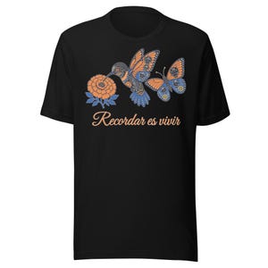 Peut inclure: Un t-shirt noir à manches courtes orné d'un motif représentant un colibri orange et bleu, un papillon et une fleur de souci. Sous le motif, le texte "Recordar es vivir" est imprimé dans une police cursive orange clair.