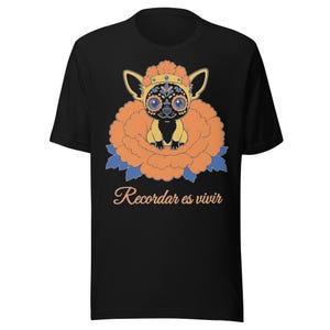 Peut inclure: Un t-shirt noir à manches courtes orné d'un motif représentant un petit chien aux marques faciales décoratives, portant une couronne et assis dans une grande fleur orange aux feuilles bleues. Le texte Recordar es vivir est imprimé en dessous.