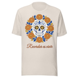 Peut inclure: Un t-shirt à manches courtes de couleur crème avec un motif central représentant un crâne en sucre blanc entouré d'une couronne circulaire de fleurs de souci orange et de feuilles bleues, avec le texte "Recordar es vivir" en brun.