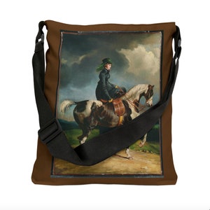 Puede incluir: Una bolsa de lona marr&oacute;n con una correa negra ajustable. La bolsa presenta una imagen impresa de una mujer montando a caballo en un campo.