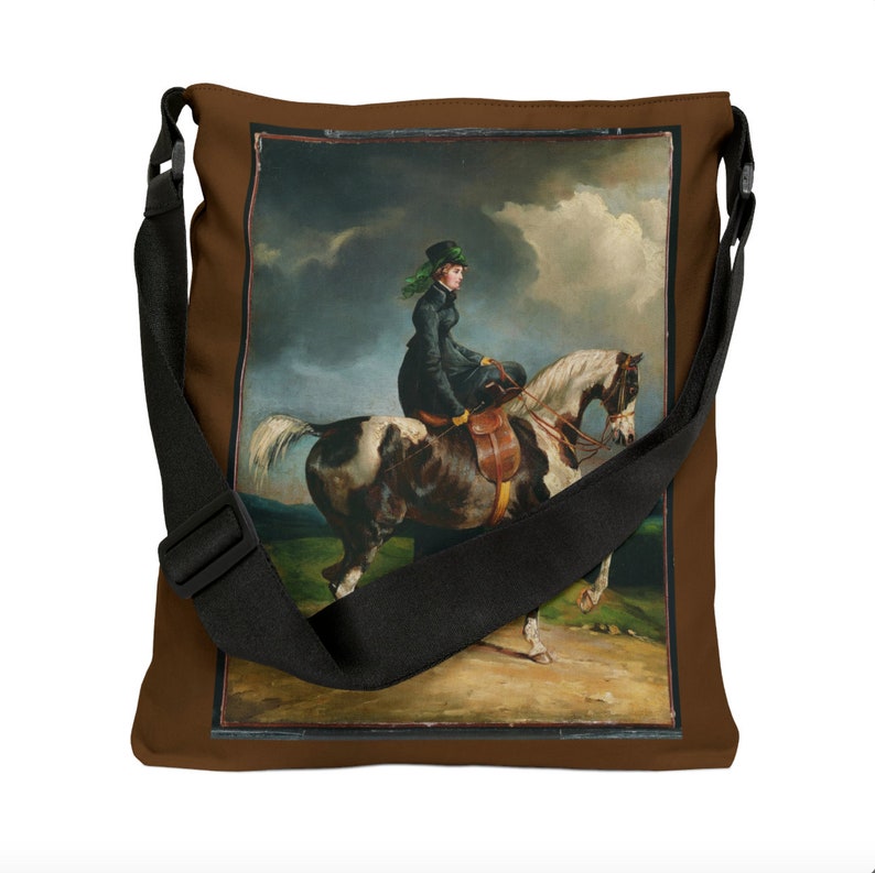 Puede incluir: Una bolsa de lona marr&oacute;n con una correa negra ajustable. La bolsa presenta una imagen impresa de una mujer montando a caballo en un campo.