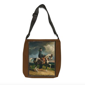 Puede incluir: Una bolsa de lona marr&oacute;n con una correa ajustable negra. La bolsa presenta una imagen impresa de una mujer montando un caballo blanco y marr&oacute;n en un campo.