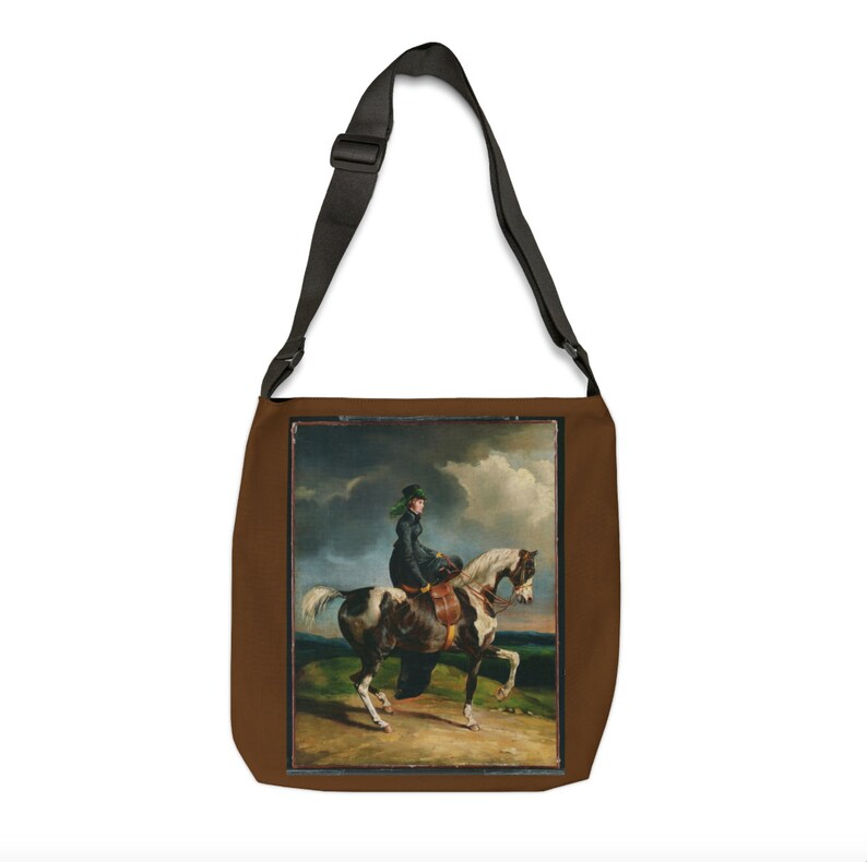 Puede incluir: Una bolsa de lona marr&oacute;n con una correa ajustable negra. La bolsa presenta una imagen impresa de una mujer montando un caballo blanco y marr&oacute;n en un campo.