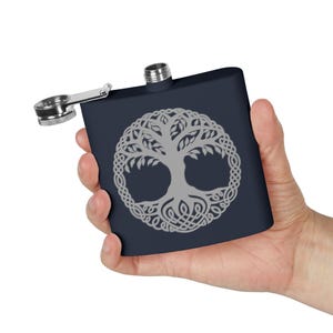 Pode incluir: Uma garrafa de bolso azul marinho com um desenho prateado da árvore da vida. A garrafa de bolso tem uma tampa articulada e uma tampa de rosca prateada. O desenho da árvore da vida é uma ilustração detalhada de inspiração celta.