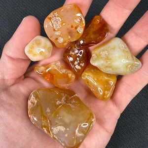 May include: A selection of polished agate stones in shades of orange, brown, and cream. The stones vary in size and shape, with some displaying unique patterns and textures. The gemstones are held in an open hand against a dark background.