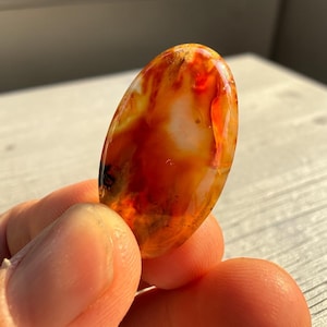 May include: Oval-shaped polished gemstone with a vibrant blend of orange, red, and white hues. The stone's surface is smooth and translucent, showcasing natural patterns. The gemstone is held between fingers, highlighting its size and shape.