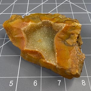 May include: A raw, amber-coloured agate specimen featuring a central druzy cavity with sparkling, light-coloured crystals. The stone displays a smooth, waxy texture on its outer edges and rests on a grid mat measuring approximately 7.5 cm wide.
