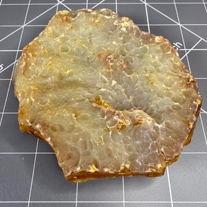May include: A translucent, irregularly shaped amber-colored stone slab with a textured, pitted surface. The stone is positioned on a dark gray cutting mat with white grid lines, showing a width of approximately 3 inches across the surface.