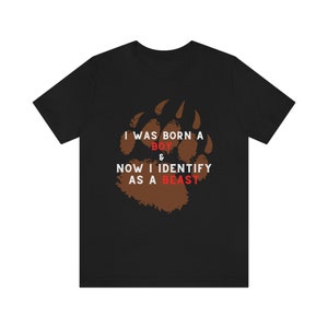 Puede incluir: Camiseta negra con un estampado de huella de pata de oso marrón. El texto "I was born a boy & now I identify as a beast" está impreso en la camiseta.