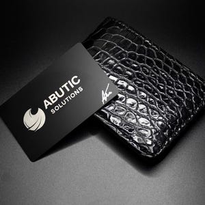 May include: A black business card with the text "ABUTIC SOLUTIONS" and a logo rests on a black, textured wallet. The wallet has a reptile skin pattern. The background is black.