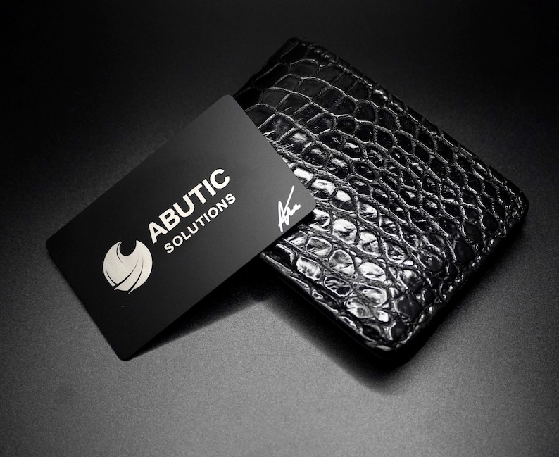 May include: A black business card with the text "ABUTIC SOLUTIONS" and a logo rests on a black, textured wallet. The wallet has a reptile skin pattern. The background is black.
