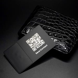 May include: A black business card with a QR code and the text "Save Contact" is displayed in front of a black leather wallet with a crocodile skin texture. The card has a rectangular design with a smooth, matte finish.