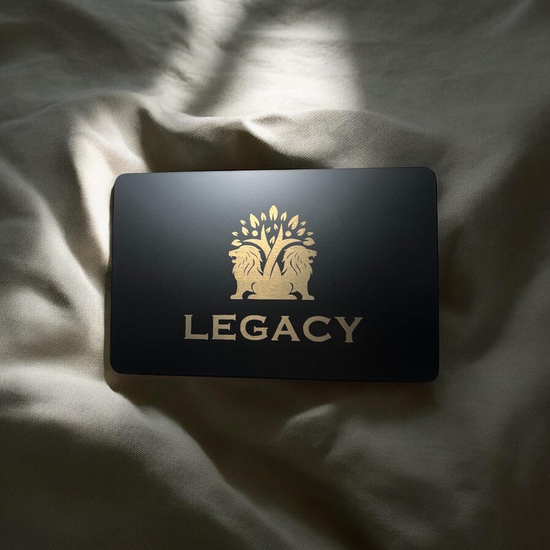 May include: A black rectangular card with gold lettering and a gold emblem. The emblem features two lions flanking a tree. The word "LEGACY" is printed in gold below the emblem. The card is resting on a light-colored, textured surface.