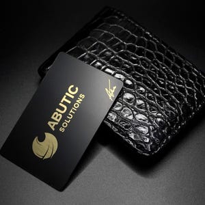 May include: A black leather wallet with a crocodile skin texture. A black business card with gold lettering that reads "ABUTIC SOLUTIONS" and a gold logo is placed on top of the wallet.