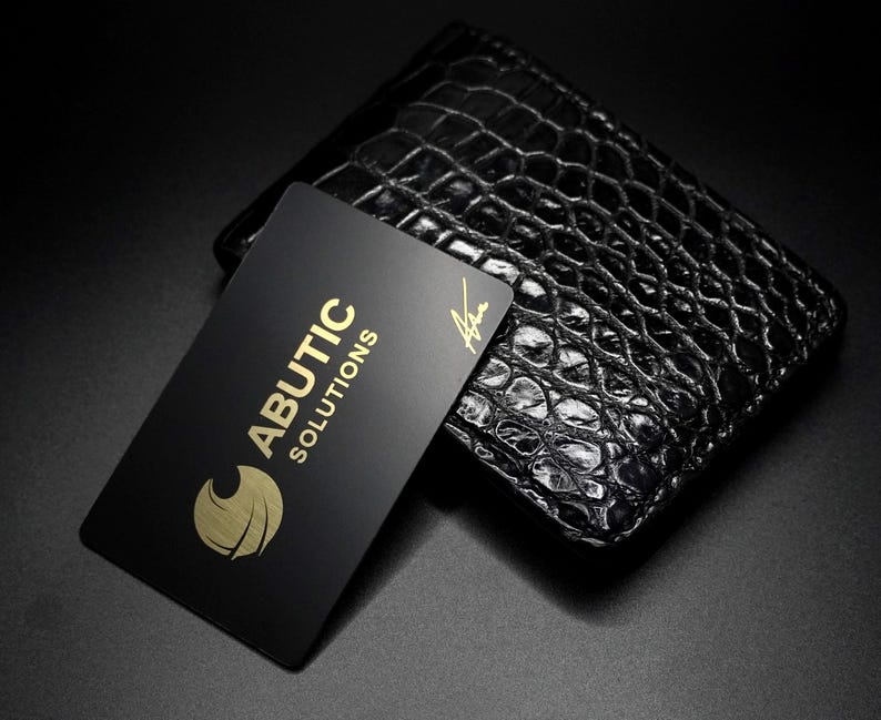 May include: A black leather wallet with a crocodile skin texture. A black business card with gold lettering that reads "ABUTIC SOLUTIONS" and a gold logo is placed on top of the wallet.