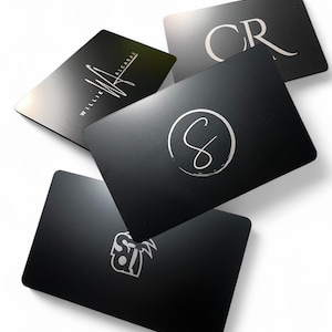May include: A collection of five black rectangular cards with silver text and logos. The cards feature various designs, including initials, a signature, and a stylized logo. The cards are arranged at different angles against a white background.