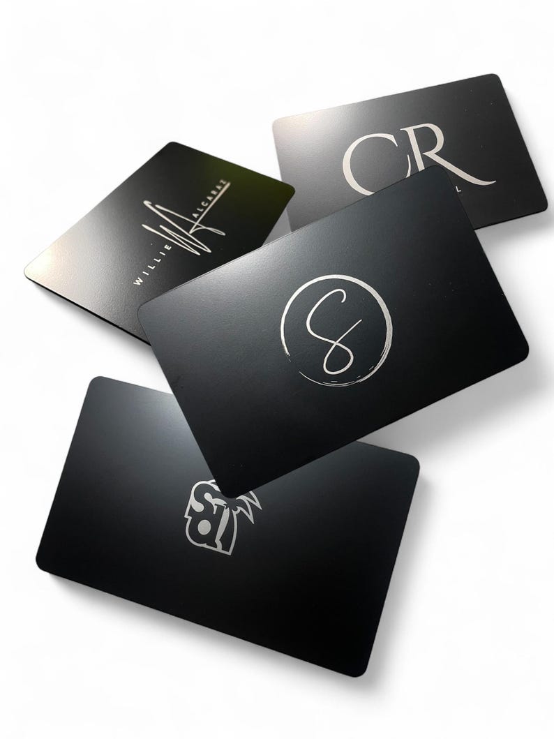 May include: A collection of five black rectangular cards with silver text and logos. The cards feature various designs, including initials, a signature, and a stylized logo. The cards are arranged at different angles against a white background.