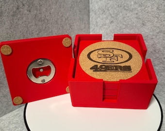 Puede incluir: Juego de posavasos rojos con un abridor de botellas a juego. Los posavasos son de corcho y presentan el logotipo de los 49ers. El abridor de botellas está integrado en un cuadrado rojo. El juego está sobre una superficie blanca.