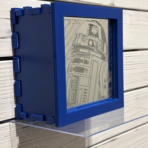 Könnte beinhalten: Ein blauer 3D-gedruckter Rahmen mit einer transparenten Acrylglasscheibe. Der Rahmen enthält ein Schwarzweißbild des Star Wars-Charakters R2-D2.