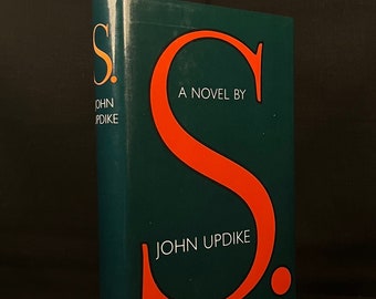 May include: A hardcover book titled "S." by John Updike. The book has a dark teal cover with a large orange "S" and the author's name in orange. The book is standing upright against a dark background.