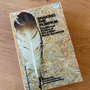 May include: A paperback book titled Shaking the Pumpkin: Traditional Poetry of the Indian North Americas, edited by Jerome Rothenberg. The cover features a large feather illustration over a marbled gold and white background with a $6.95 price tag.