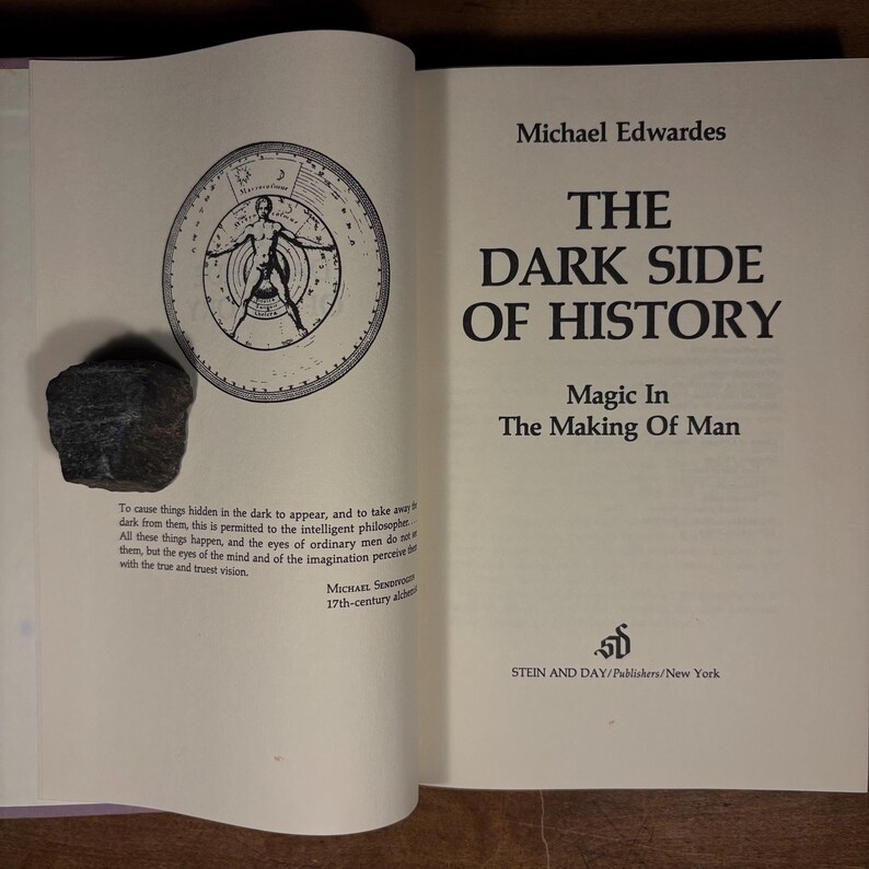 May include: Open book with the title "THE DARK SIDE OF HISTORY" by Michael Edwardes. The book is open to a page with text and an illustration of a figure inside a circle. A dark gray rock sits on the open page.