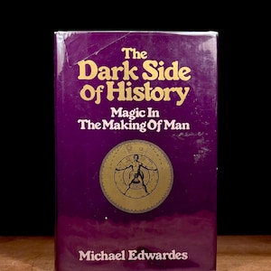 May include: A vintage book titled "The Dark Side of History" by Michael Edwardes. The cover is a deep purple with gold lettering and a circular illustration. The book's subtitle reads "Magic In The Making Of Man."