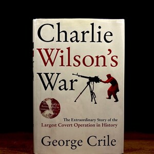 May include: A white book cover with the title "Charlie Wilson's War" in black and red lettering. The cover features a silhouette of a person operating a machine gun. The text "The Extraordinary Story of the Largest Covert Operation in History" is printed below the image. The author's name, "George Crile", is printed in black at the bottom of the cover.