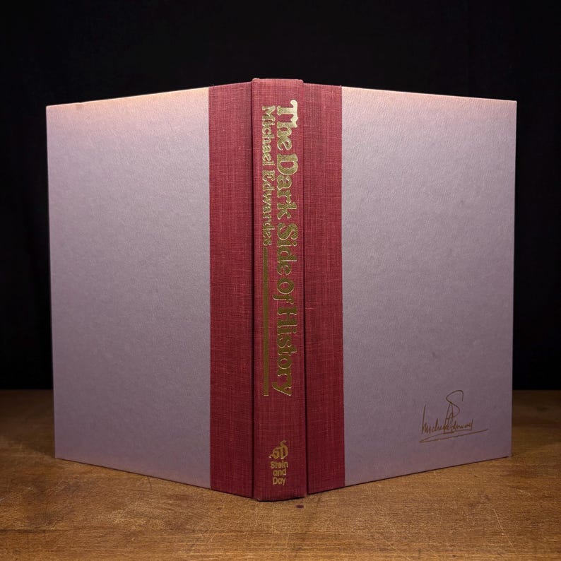May include: A hardcover book titled "The Dark Side of History" by Michael Edwards. The spine is burgundy with gold lettering. The book's cover is a pale lavender color. The book is displayed open, revealing the spine and part of the cover.