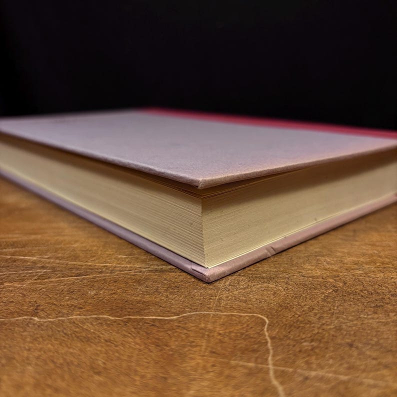 May include: A hardcover book with a light purple cover and a red spine. The pages are yellowed, and the book is resting on a wooden surface. The background is black.