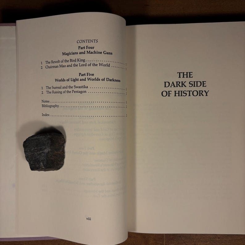 May include: An open book with the title "THE DARK SIDE OF HISTORY" on the right page. The left page displays the table of contents, including "Magicians and Machine Guns" and "Worlds of Light and Worlds of Darkness." A dark gray rock sits on the left page.