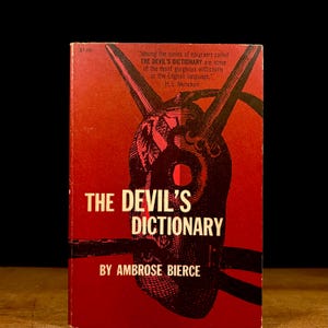 May include: A vintage copy of "The Devil's Dictionary" by Ambrose Bierce. The book has a red cover with a black illustration of a horned mask. The title and author's name are in white. The cover also includes a quote.