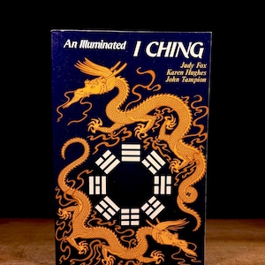 May include: A hardcover book titled "An Illuminated I Ching" with a dark blue cover. The cover features gold dragons and geometric symbols. The authors' names are listed on the right side of the cover.
