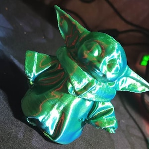Puede incluir: Una figura impresa en 3D de un personaje verde con orejas grandes y una túnica con capucha. La figura tiene un acabado brillante e iridiscente.