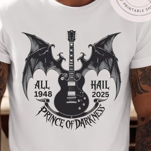 Könnte beinhalten: Weißes T-Shirt mit einem schwarzen Gitarrendesign, Fledermausflügeln und dem Text "ALL 1948 HAIL 2025 PRINCE OF DARKNESS". Die Grafik ist mittig auf dem T-Shirt platziert.