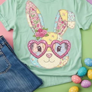 Op de afbeelding: Een lichtgroen t-shirt met een patchwork konijnengezicht, hartvormige bril en bloemmotieven. Het konijn heeft roze en gele oren en een klein boeket roze rozen. Een blauwe spijkerbroek en paaseieren staan ook op de afbeelding.