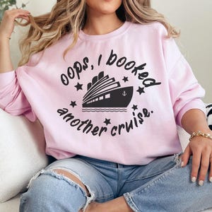 Puede incluir: Sudadera rosa con el texto "oops, I booked another cruise." y una silueta negra de un crucero.