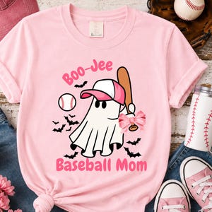 May include: A light pink t-shirt featuring a graphic of a white ghost wearing a pink and white baseball cap. The ghost holds a brown baseball bat with a pink bow. The text reads Boo-Jee Baseball Mom, surrounded by small black bats and a baseball.