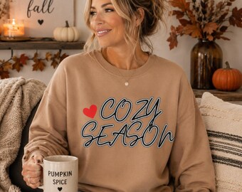 May include: A tan crewneck sweatshirt featuring the text "COZY SEASON" in black script with a red heart icon. The sweatshirt is paired with a light-coloured ceramic mug that has the text "PUMPKIN SPICE" and a small black heart printed on it.