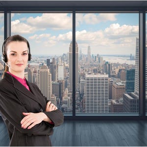 May include: A woman wearing a headset and a black blazer, standing in front of a large window with a city skyline view. The woman has her arms crossed. The background features a cityscape with tall buildings and a cloudy sky.