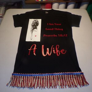 May include: Black t-shirt with a red graphic that says "I Am Your Good Thing Proverbs 18:22 A Wife". The graphic also includes a black and white illustration of a woman's face. The shirt has a red, white, and blue fringe trim.