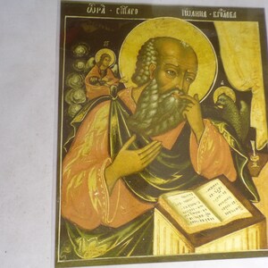 May include: A religious icon depicting Saint John the Evangelist, with a halo and a book in his hand. The icon is painted in a traditional style with gold accents and a dark background. The text "Θερα - Σιγαλο" and "Ιωαννης - Εγκομεδα" is visible at the top of the icon.